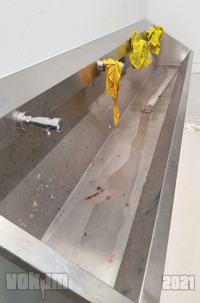 Big metal sink with four auto stop taps. There are yellow cleaning cloths draped on the last three taps. Rest of the photo are white walls and floor. The sink is a bit stained with paint from cleaning brushes.